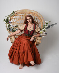 Full length portrait of beautiful red haired woman wearing a medieval maiden, fortune teller costume.  Sitting pose, with gestural hands reaching out. isolated on studio background.