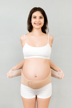 Portrait of pregnant woman in underwear dressing maternity belt against pain in the back at gray background with copy space. Orthopedic abdominal support belt concept