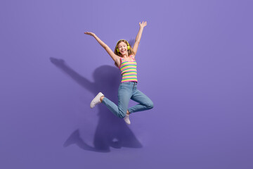 Fototapeta na wymiar Full size photo of pretty teenager blonde girl jump raise hands excited wear trendy striped outfit isolated on purple color background