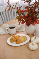 Autumn home interior. Two croissans on the plate, glass of tea, vase with autumn leaves on wooden table near the window.