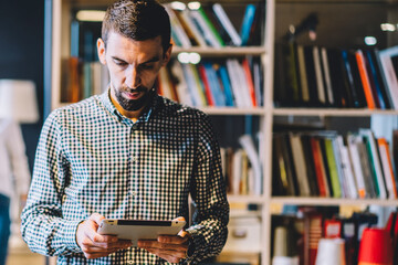 Calm pensive businessman using digital tablet while colleagues walking past and talking to each other in open space modern office