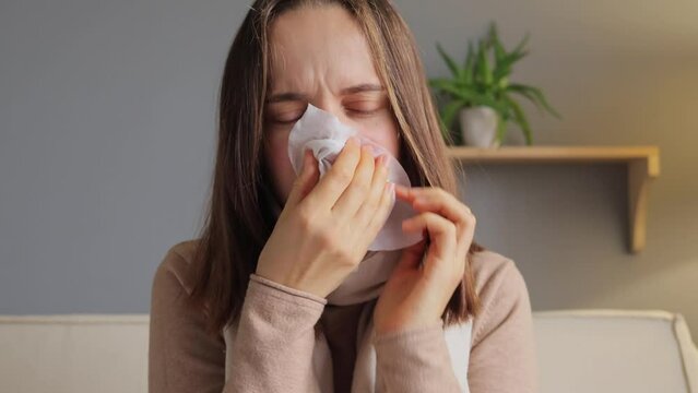 Flu symptom. Sick woman. Seasonal allergy. Unwell sad brown haired woman covid infection blowing nose in tissue suffering runny nose in home interior.