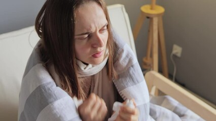 Unwell woman sitting at home wrapped in plaid shivering coughing has high temperature staying at home during quarantine while having flu.