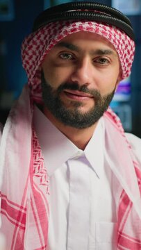 Vertical video POV of cheerful Muslim content creator dressed in traditional clothing filming himself. Smiling Arab man with thobe and headscarf waving towards camera, doing online vlogging