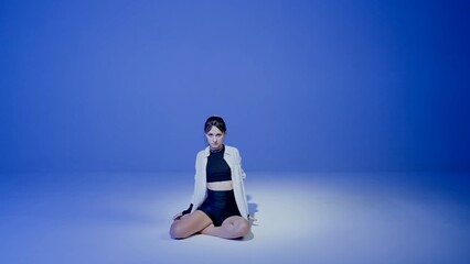 Young woman wearing a top, shorts and a shirt performing contemporary dance under a spotlight in the studio. Neon blue color scheme, ombre, gradient background. Full length.