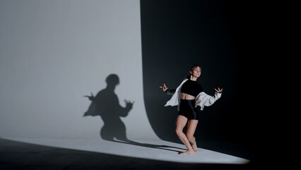 Young woman wearing a top, shorts and a shirt performing contemporary dance under a spotlight in the studio. Contrasting black falling shadow on the background. Full length.