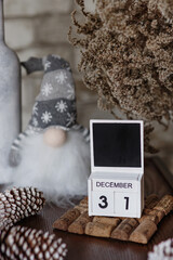 New year winter seasonal cozy decor: wooden calendar with date December 31, Christmas dwarf toy in knitted cap, cones, dried branches, cork board, bricks background, vertical festive still life