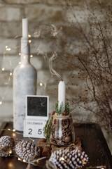Christmas festive cozy vertical decorative composition: white bottle with candle, extinguished candle with curved smoke, wooden calendar with date December 25, cones, lights garland, bricks background
