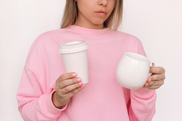 Young blonde woman in pink sweetshirt holding white paper cup with lid and glass cup choosing which...