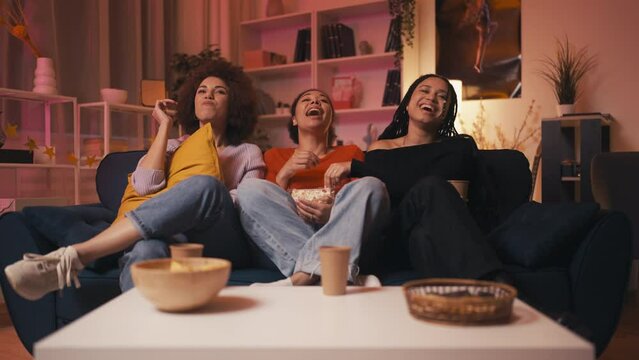 Happy black female friends watching comedy at home, eating popcorn and laughing