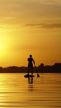 Vertical Screen: Female silhouette on SUP board in beautiful landscape at sunset. Woman paddling, her reflection on water with ripples. Outdoor activity and healthy lifestyle. Concept of fitness