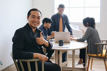 Smiling confident young Asian businessman looking at camera and showing thumbs up in office at team...
