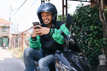 Male online motorcycle taxi driver using smartphone and sitting on scooter while waiting for an...