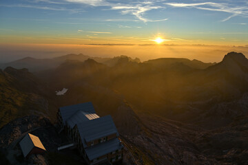 Fototapeta premium Sonnenaufgang in den Bergen mit einer Berghütte im Vordergrund und Silhouetten von Gipfel im Morgendunst.