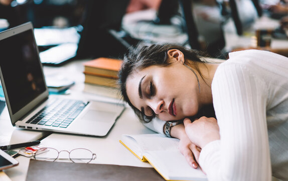 Exhausted female student sleeping on table with laptop and copybook at library