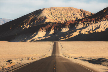 Fototapeta na wymiar Dramatic golden light on an empty desert road straight ahead through the rugged terrain of the badlands landscape in Death Valley Park National Park, USA.