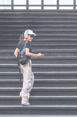 Dark-skinned Asian female tourist in casual clothing style using mobile phone while walking up the stairs at outdoor elevated view point, full length and vertical frame