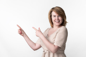European blonde woman mature adult dressed in beige dress with belt tied in front looking at camera, standing on white background. Female points fingers upward at something interesting. Copy space