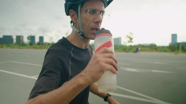 Medium shot of male cyclist in helmet standing with bike on road and drinking water from bottle while having break