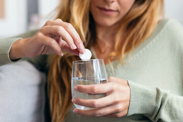 Unhealthy young woman with headache putting a pill in a glass of water sitting on sofa at home.