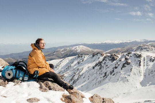 Portrait of a man in the mountains: a man sits on a peak with a backpack behind him against a mountain backdrop. High-quality photo for website design, postcards, banners, and travel product advertisi