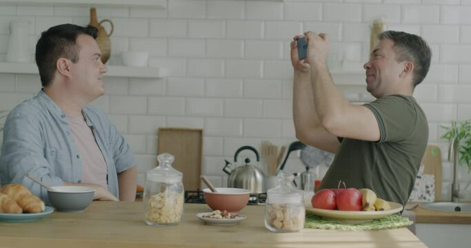 Playful gay man posing for camera having fun while boyfriend taking photo with smartphone at kitchen table. Photography and same sex relationship concept.