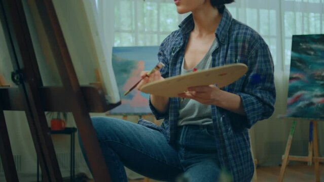 A woman is rustling through many different types of brushes while painting something astonishing on a large canvas that is held up by an easel