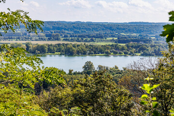 Maas River seen through foliage of lush green trees on Caestert Plateau, sailboat and boat sailing, Dutch countryside in background, border between Holland and Belgium, sunny day in Riemst, Belgium