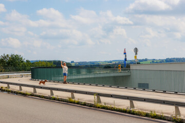Rural road and pedestrian path and bicycles at Lanaye locks, smiling senior female tourist standing with her dog next to a metal fence waving with raised hand, sunny summer day in Ternaaien, Belgium
