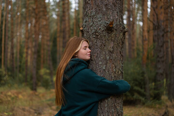 A young red-haired woman hugs a tree in a pine forest and smiles happily