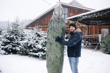 Happy handsome bearded man holds a packed live Christmas tree in outdoor market. Winter snowy day