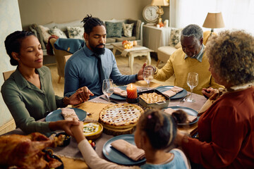 Extended black family saying grace during Thanksgiving meal at dining table.