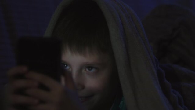 Young caucasian boy using phone in the dark, covered with blanket. Toddler playing games late at night. Cute little boy sitting in bad and watching cartoons. Kid having fun.
