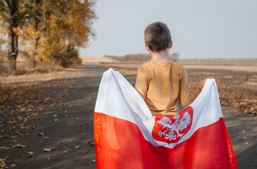 A handsome little boy behind his back holds the flag of Poland in his hands. Child standing on road with national flag on nature. Concept of national holidays and patriotic events. Independence Day