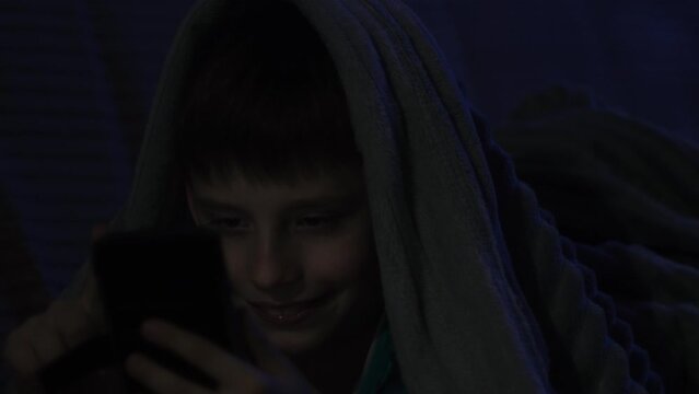 Young caucasian boy using phone in the dark, covered with blanket. Toddler playing games late at night. Cute little boy sitting in bad and watching cartoons. Kid having fun.
