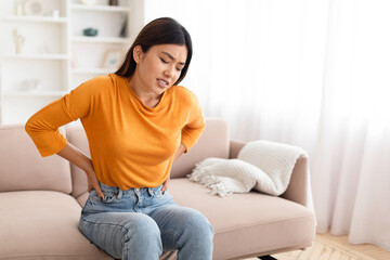 Unhappy asian woman sitting on couch, touching back, have pain