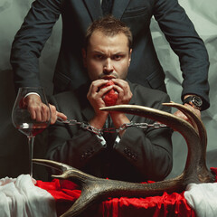 Devil concept. Portrait of captured criminal eating fresh heart. Policeman holding hands of cannibal with metal chain. Decadent accessories. Cinematic style. Studio shot