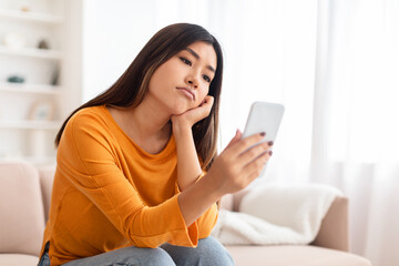 Bored asian young lady using mobile phone at home