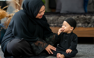 Muslim mother wearing traditional black dress and headscarf, feeding food to baby boy, eating with...