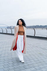 Woman in a coat outdoors. Model in outerwear. A brunette with curly hair in a long beige coat walks along the river embankment. Beautiful girl in a fashionable casual coat. spring-autumn collection