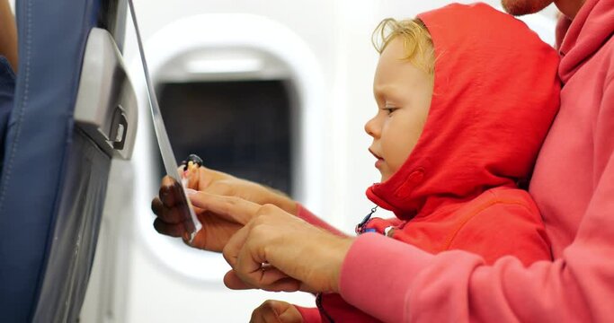 A baby passenger and his father enjoy in-flight bonding, with the young child's curiosity piqued by the captivating images in the airplane's safety instructions during their journey.