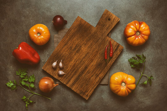 Culinary background. Empty wooden cutting board, fresh vegetables, greens and spices on a dark rustic background. Top view, flat lay, copy space. Toned image