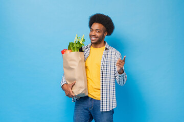 Handsome friendly smiling Afro African American man holding paper shopping bag full of groceries in light blue isolated studio shot background - obrazy, fototapety, plakaty