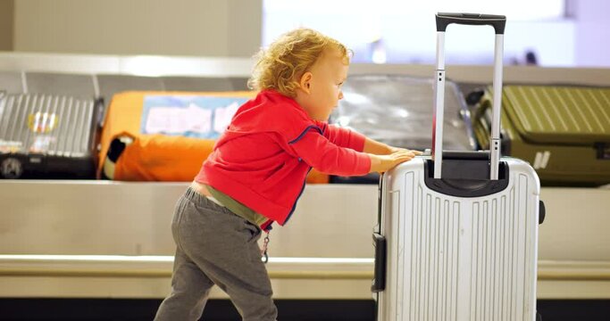 Confident toddler, self-sufficient and adorable, pushes small suitcase through airport. Slow motion trucking shot at baggage claim hall, young child walks against moving conveyor belt with cases.