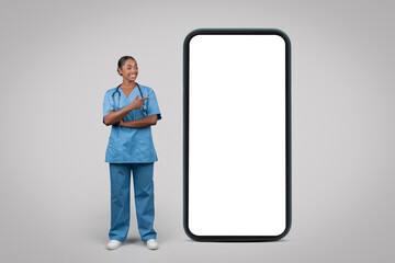 Cheerful black millennial woman nurse, surgeon in blue uniform point finger at big phone with empty screen