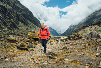 Woman in sunglasses with backpack and trekking poles dressed red softshell jacket hiking during...