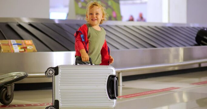 Cheerful toddler plays in airport baggage claim area, lifting, dropping, and flipping small suitcase in slow motion while waiting for his luggage. Selective focus, blurred conveyor belt with bags
