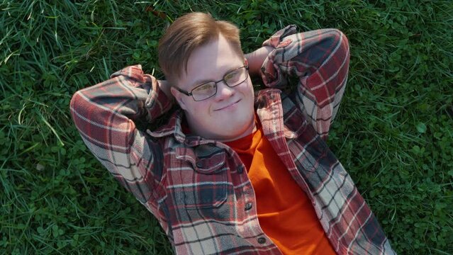 Portrait of young man with down syndrome in checkered shirt laying on the grass outdoors in the public park. Man with trisomy 21 illness. Disabled guy having a rest
