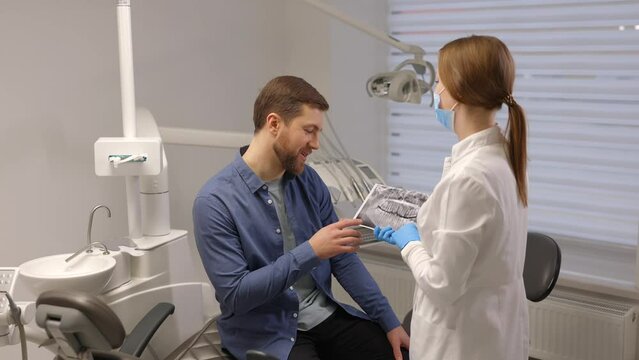 Young attractive man visiting dentist, sitting in dental chair at modern light clinic. Young woman dentist holding x ray image