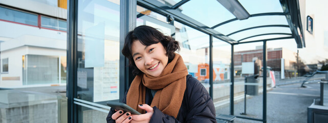 Cute smiling asian girl standing on bus stop, holding smartphone, wearing winter jacket and scarf....